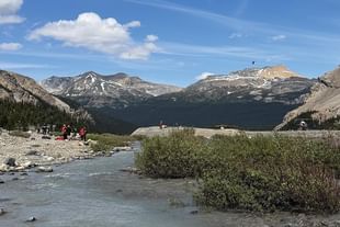 Mountain landscape with a flowing stream, evergreen trees, and snow-capped peaks under a blue sky with white clouds and hikers in the distance