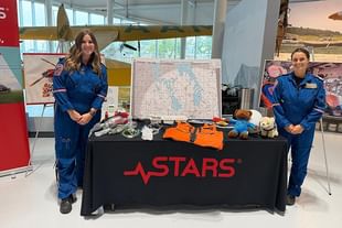 Two people in blue flight suits stand at a STARS emergency medical services exhibition table displaying equipment and maps in a modern indoor venue.