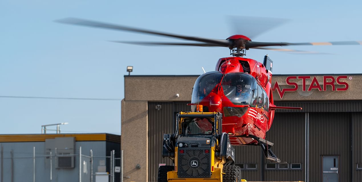 A red STARS air ambulance helicopter begins taking off from it's landing pallet outside the STARS Winnipeg base against a blue sky and the hangar visible in the background.
