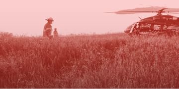 Helicopter landing in a vast grain field with a person standing nearby in a monochromatic red-toned landscape