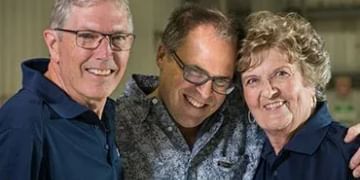 Three smiling people posing together indoors, with a man on the left wearing glasses and a dark blue shirt, a man in the center wearing a patterned shirt and glasses, and a person on the right wearing a dark blue shirt with "STARS" visible on it.