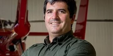 A smiling person in an olive green shirt standing in front of red industrial equipment in a garage or workshop setting.