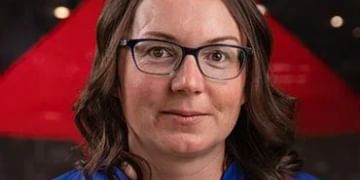 Person with long dark hair and glasses wearing a blue shirt, photographed against a red geometric background
