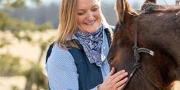 A person with blonde hair smiling while petting a brown horse outdoors in a sunny, natural setting with trees in the background.