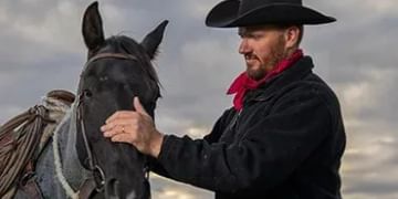 Cowboy in black hat and red bandana petting a black horse against a cloudy sky background