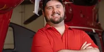 Person in red shirt and cap standing with arms crossed in front of red machinery or equipment in an industrial setting.