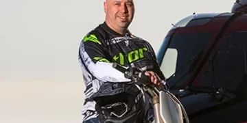 Person in black and neon green jacket standing next to a bright green motorcycle, with a red vehicle visible in the background against a clear sky.