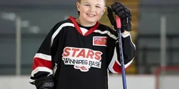 Young hockey player wearing black, red and white Winnipeg Stars jersey, holding a stick and smiling at an indoor ice rink