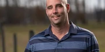 A person with a bald head wearing a blue and navy striped polo shirt, smiling at the camera outdoors with a blurred green field in the background.