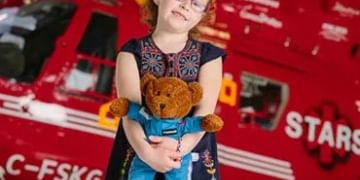 Child wearing glasses holding a teddy bear in front of a red helicopter or aircraft with registration marking C-FSKG