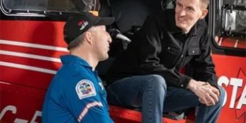 Two people sitting in a red fire truck cabin, one wearing a blue uniform with patches and a black cap, the other in dark clothing, both smiling at the camera.