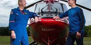 Two people in blue flight suits standing in front of a red helicopter in an outdoor field during daytime