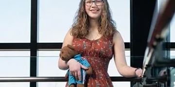Person wearing glasses and a red floral dress holding a blue object, standing in front of large windows with a city view visible in the background.
