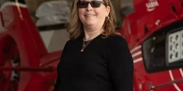 Person wearing sunglasses and black ribbed sweater standing in front of red aircraft or vehicle in hangar setting