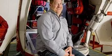 Person sitting inside a spacecraft or aircraft cabin, wearing a gray shirt and jeans, surrounded by red equipment and technical apparatus.