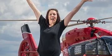 Person with arms raised in celebration standing in front of a red helicopter against a cloudy blue sky
