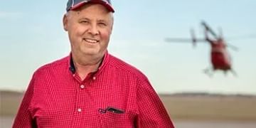 Person in red checkered shirt and cap standing outdoors with helicopter in blurred background sky