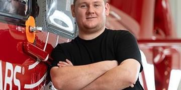 Person in black shirt standing with arms crossed in front of red vehicle or equipment with partial text visible, indoor setting with window in background