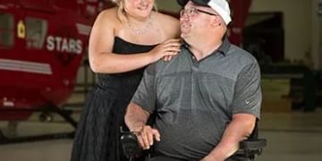Person in wheelchair wearing gray shirt and cap with another person in black dress standing beside them, both smiling in what appears to be an indoor venue with red signage
