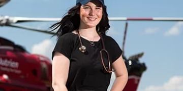 Person in black shirt and blue cap smiling at camera with red helicopter in blurred background under blue sky