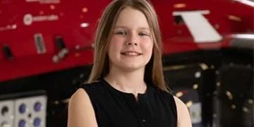 Young person with long brown hair wearing a black sleeveless top, smiling at camera with a red aircraft visible in the background.