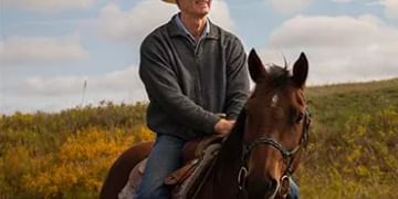 Cowboy in tan hat and dark jacket riding a brown horse across grassy prairie under cloudy sky