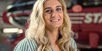 Person with long blonde wavy hair wearing a blue and white striped shirt, smiling at camera in front of a blurred garage or workshop setting