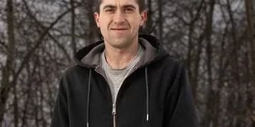 Person wearing a black zip-up hoodie and gray shirt standing outdoors with bare trees in the background during daytime.