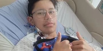 Person wearing glasses and a blue shirt lying in a hospital bed, giving a thumbs up gesture while holding medical tubing