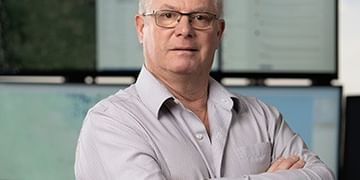 Professional headshot of a person wearing glasses and a light gray button-up shirt, standing with arms crossed in front of a modern office background with multiple screens.