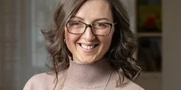 Person with shoulder-length brown hair and glasses wearing a beige turtleneck sweater, smiling at the camera in an indoor setting with bookshelves visible in the background.