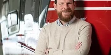 Man with beard wearing beige sweater standing with crossed arms in front of red and white striped vehicle or equipment