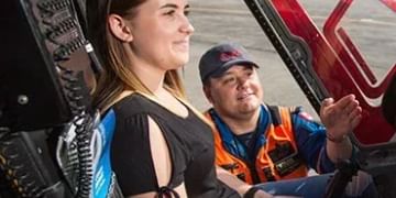 Two people sitting in what appears to be a vehicle or machinery, with one wearing a black shirt and the other wearing an orange and blue safety vest and cap.