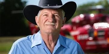 Older person wearing a black cowboy hat and light blue shirt smiling at camera with farm equipment blurred in background