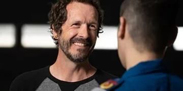 Two people conversing in front of a dark background, one wearing a black and gray shirt smiling at the other who wears a blue jacket.