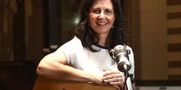 Person with long dark hair smiling at a microphone while holding an acoustic guitar in a recording studio setting