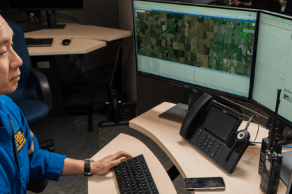 Person in blue shirt working at computer desk with dual monitors displaying aerial/satellite imagery and maps in an office environment