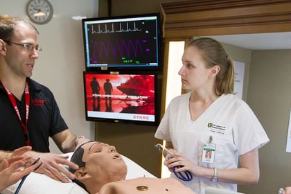 Healthcare professionals demonstrating cardiac examination techniques on a training mannequin while monitoring vital signs on displays above
