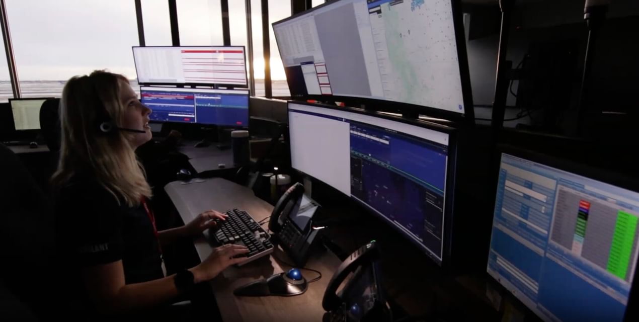 Person working at a multi-monitor workstation displaying data analytics and software interfaces, with large windows overlooking a waterfront in the background.