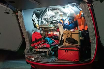 Vehicle cargo area packed with outdoor equipment including red bags, climbing gear, and supplies, with two people wearing safety vests and helmets visible in the bright interior.