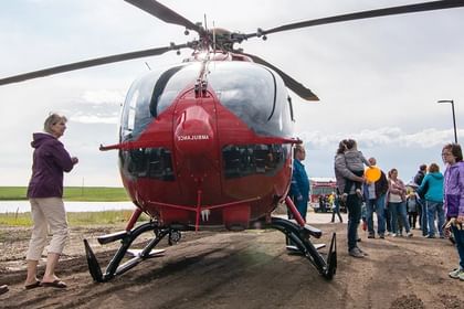 Red and white helicopter on ground surrounded by visitors and staff in outdoor setting with fields and water visible in background