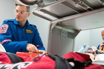 Two recovery personnel in blue flight suits examine equipment inside a spacecraft cabin, with red and white parachutes visible in the foreground.
