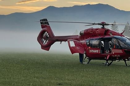 Red STARS helicopter landed in misty field with forested mountains and evergreen trees in background at sunrise or sunset.