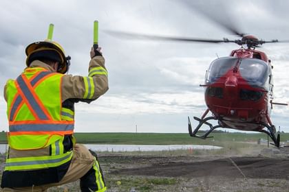 Ground crew member in high-visibility vest and hard hat signals to red helicopter landing on rural airfield with green fields and buildings visible in background