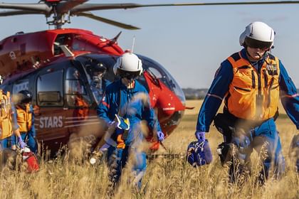 Emergency medical personnel in blue and orange gear walking from a red and blue helicopter landed in a dry grassland field under clear sky.