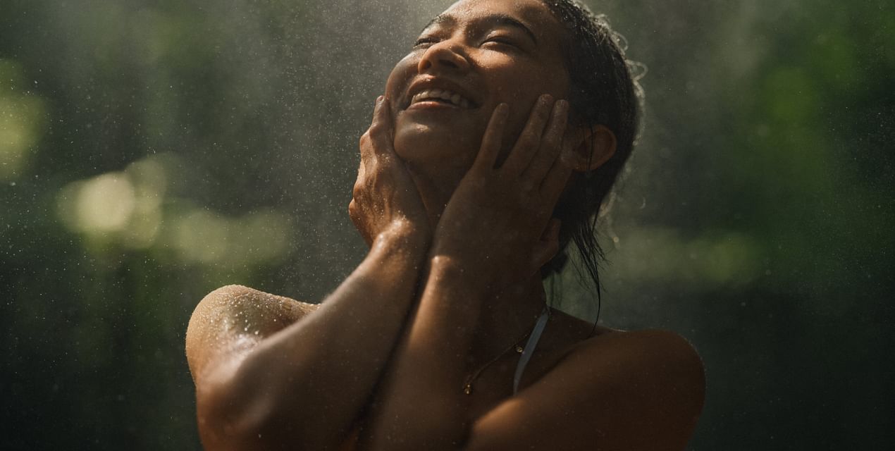 Person with wet skin and long dark hair tilting head back with eyes closed and a smile, standing in heavy rain with a blurred green natural background