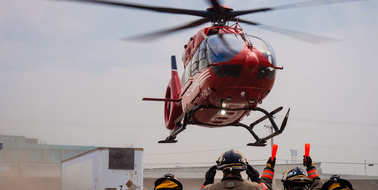 A firefighter helps land a STARS air ambulance at an industrial site, while other firefighters observe the landing.