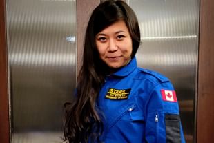 Dr. Katie Lin wears a blue STARS flight suit with a Canadian flag patch, standing in front of a textured metal elevator door, smiling at the camera.