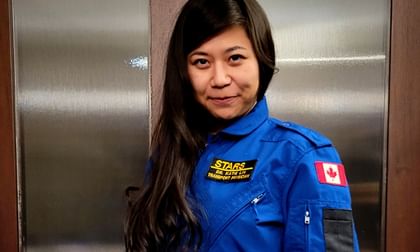 Dr. Katie Lin wears a blue STARS flight suit with a Canadian flag patch, standing in front of a textured metal elevator door, smiling at the camera.