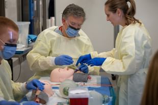STARS transport physician Dr. Jen Nicol directs STARS President and CEO Dr. John Froh during medical training at a STARS seminar. Medical professionals in protective gowns and blue gloves performing a procedure on a mannequin head during a training session in a clinical setting.